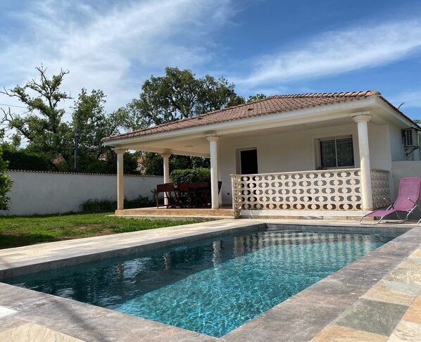 A swimming pool with a tiled deck, a covered patio with a pergola, and a white wall with a window.