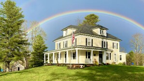 A two-story house with a porch, a flag, and a rainbow in the background.