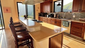 A modern kitchen with wooden cabinets, a large island with bar stools, and a view of the outdoors. - Villa HERMOSA a BEAUTIFUL St. John Villa - Pool - Ocean and Mountain Views (St John)
