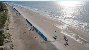 Eine Vogelperspektive zeigt einen Strand mit blauen Sonnenschirmen und Liegestühlen, eine sandige Küstenlinie und das Meer im Hintergrund.