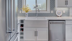 Eine moderne Küche mit weißen Schränken, einem Edelstahl-Kühlschrank, einem Geschirrspüler und einem Fenster mit Jalousien.