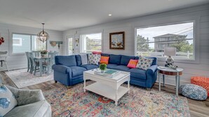 Ein geräumiges Wohnzimmer mit einem blauen Ecksofa, einem weißen Couchtisch und einem farbenfrohen Teppich. Es gibt einen Essbereich mit Tisch und Stühlen, und ein Fenster mit Blick auf ein Haus.