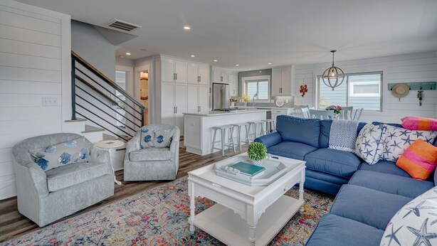 Ein modernes Wohnzimmer mit einem blauen Sofa, grauen Sessel und einem weißen Couchtisch. Ein bunter Teppich und eine Treppe, die in eine obere Ebene führt, komplettieren das Bild.
