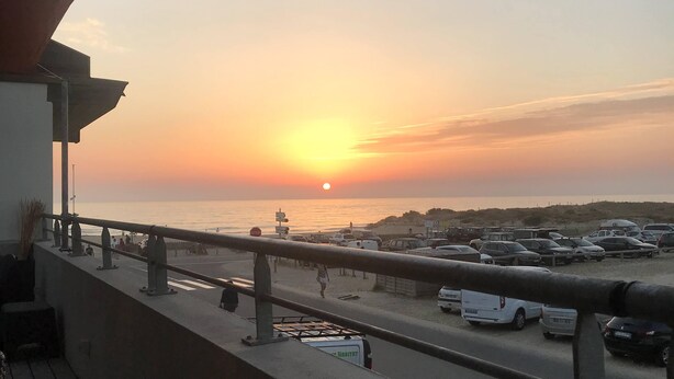 A sunset view from a balcony overlooking a beach with parked cars and a parking lot.