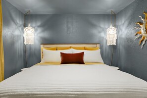 Een bed met een houten hoofdbord, wit beddengoed en een enkel bruin kussen. Aan weerszijden van het bed hangen twee lampen.