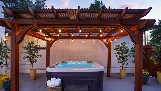 Un bain à remous sous une pergola en bois, illuminé de guirlandes lumineuses, entouré de plantes en pot et bordé d’un mur de briques.