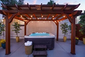 Een hottub onder een houten pergola met lichtjes, omringd door plantenpotten en een bakstenen muur.