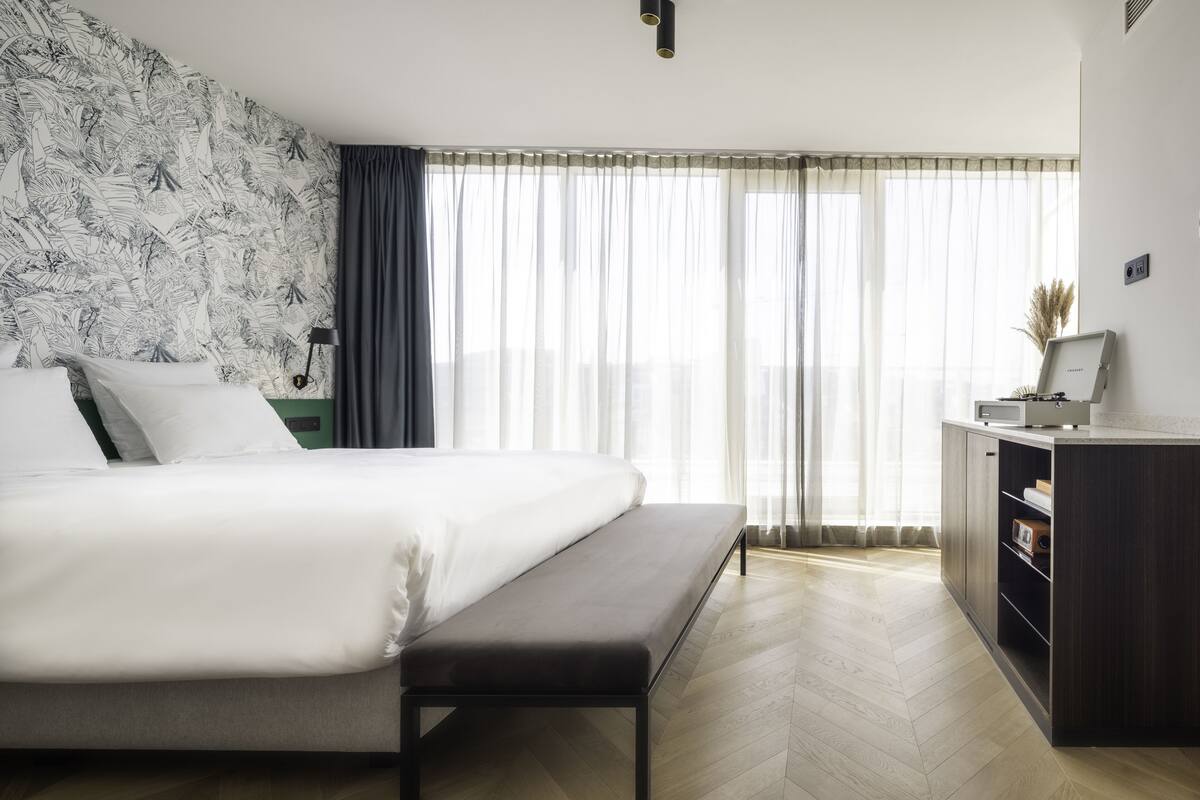 Et moderne hotelværelse med en stor seng, en bænk, et natbord, et skrivebord med lampe og udsigt over byen gennem lette gardiner.