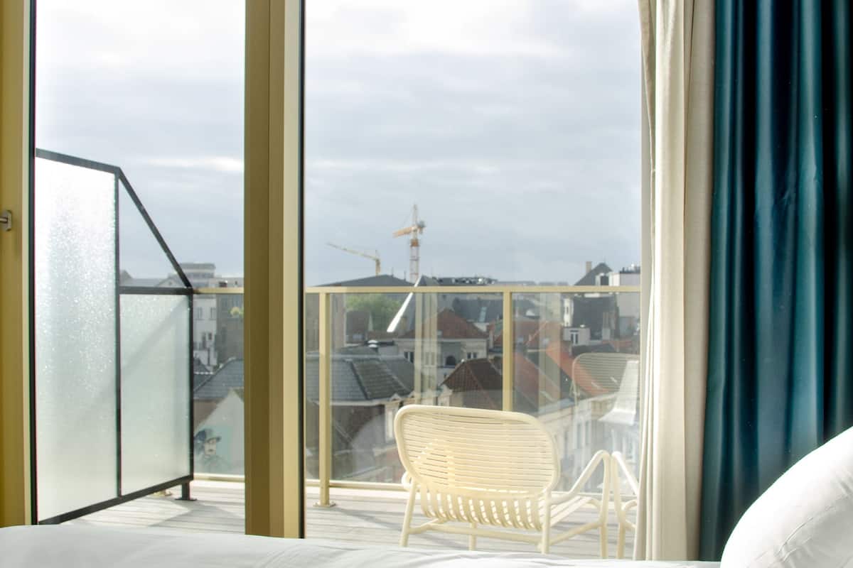 Et hotelværelse med en stor seng, en hvid stol og en balkon med udsigt over bybilledet.