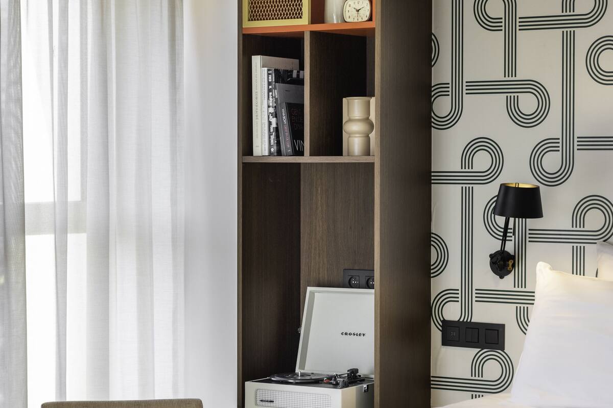 Et moderne hotelværelse med en seng, et skrivebord med en bærbar computer, en reol og en dekorativ vægpanel.