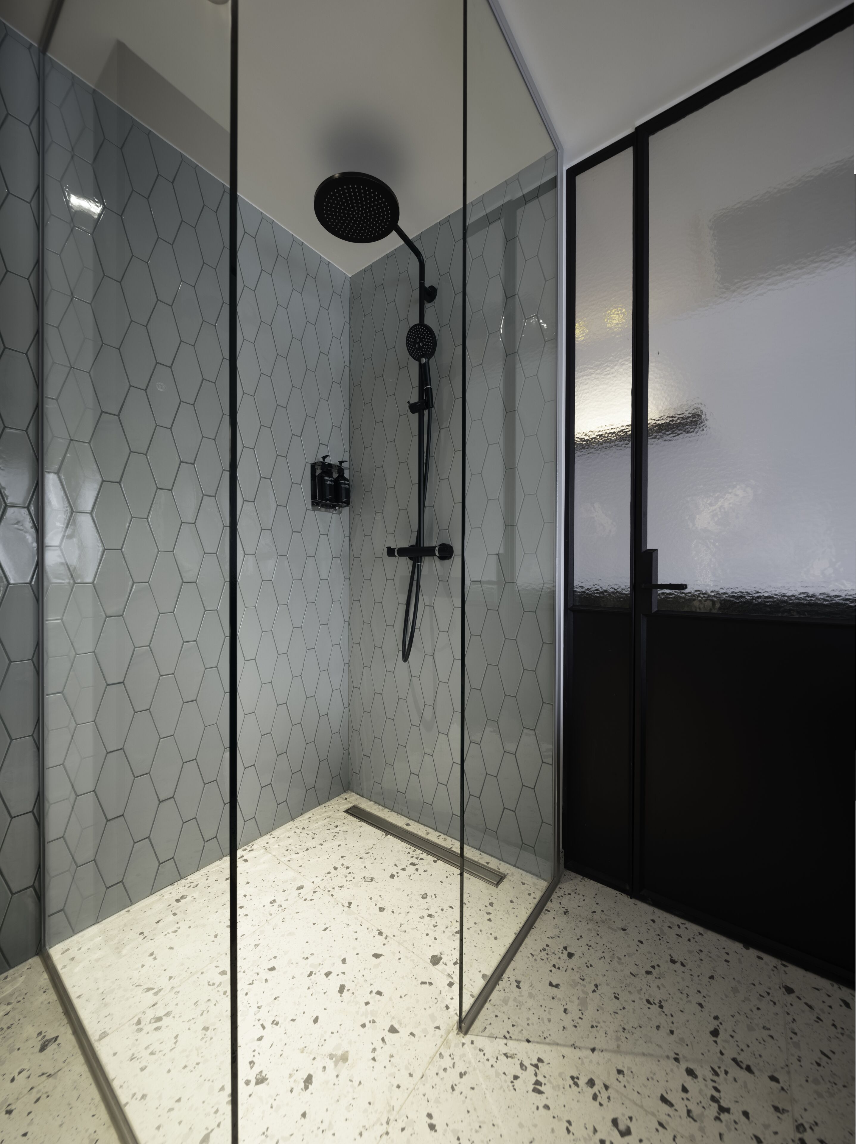 A modern shower with a glass door, hexagonal tile walls, and a speckled floor.