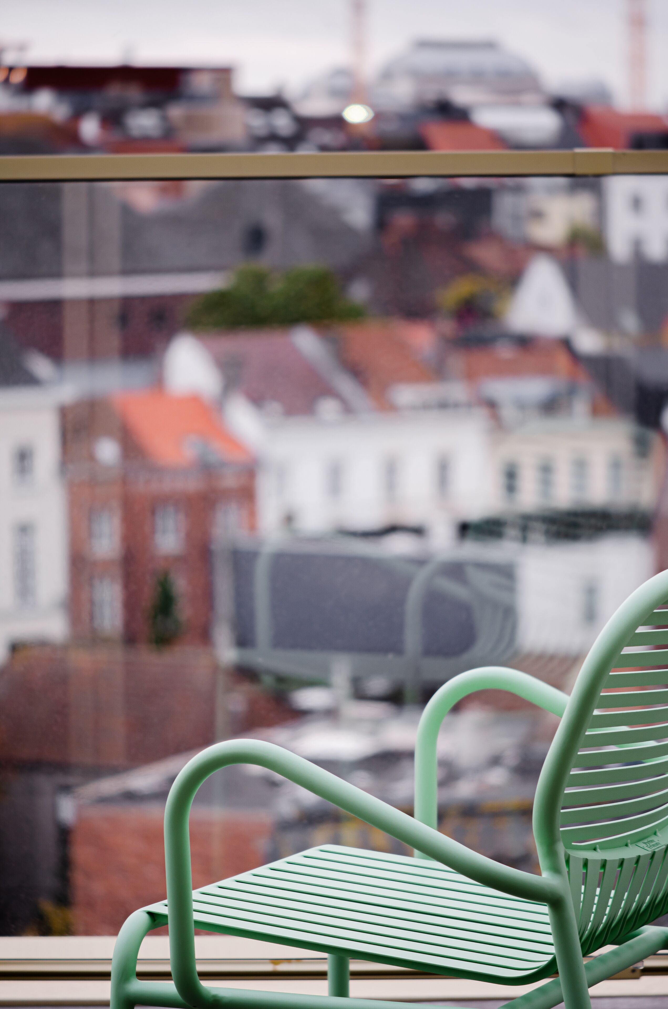Une chaise verte est placée devant une fenêtre donnant sur un paysage urbain avec des bâtiments et une structure surmontée d’un toit rouge.