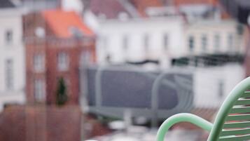 Une chaise verte est placée devant une fenêtre donnant sur un paysage urbain avec des bâtiments et une structure surmontée d’un toit rouge.