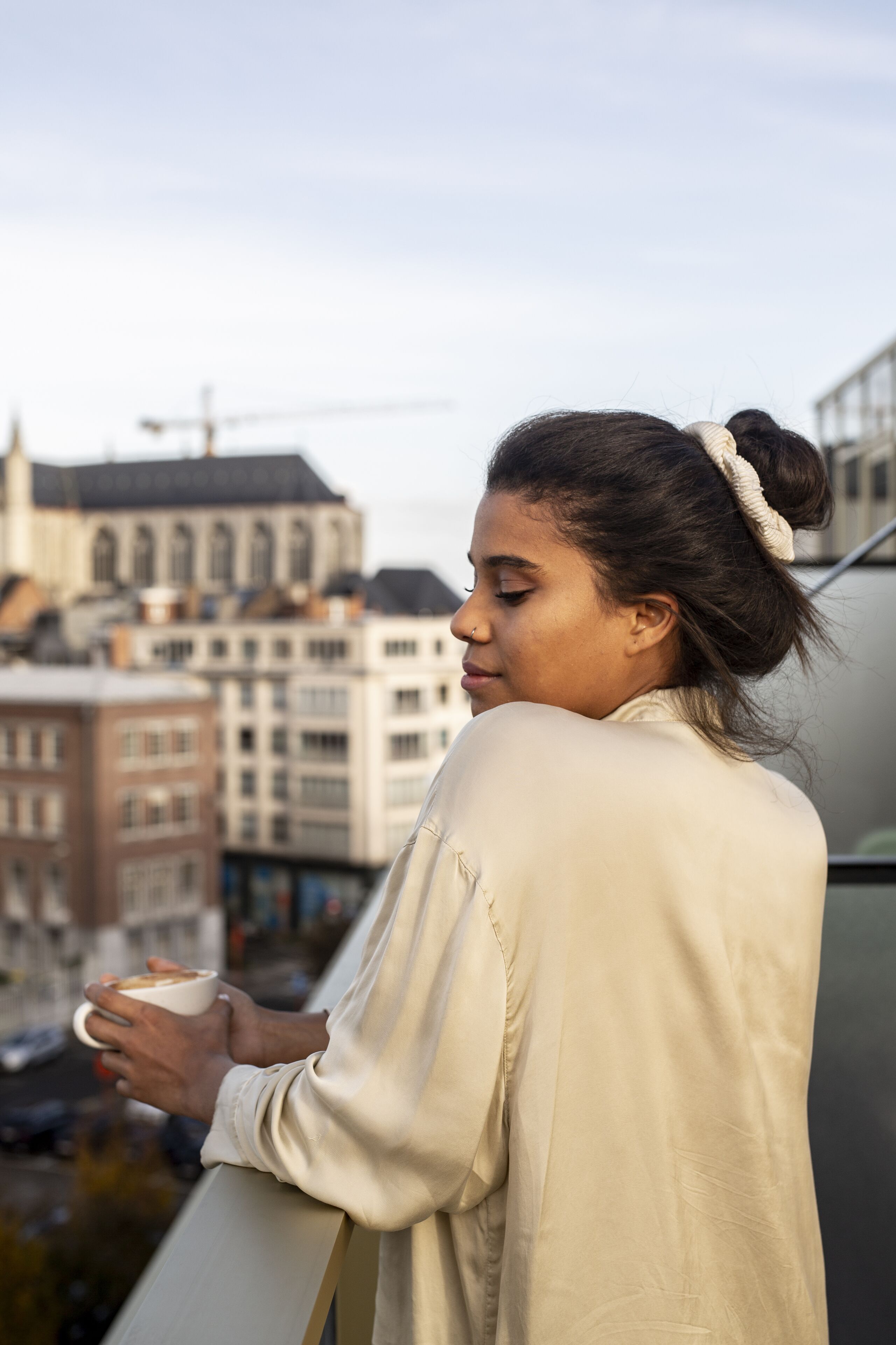 A person holding a cup, standing on a balcony with a cityscape in the background.
