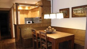 A wooden dining table with four chairs and a bowl of round objects. A kitchen area with wooden cabinets and a window is visible in the background. - Bellver Center (Bellver de Cerdaña)