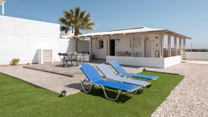 A modern outdoor area with two blue lounge chairs and a table, a white building with a covered patio, and a palm tree. - GK in Santorini Home ART (Santorini)