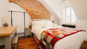 Un dormitorio con techo inclinado, pared de ladrillo, cama con ropa blanca, escritorio de madera y radiador.