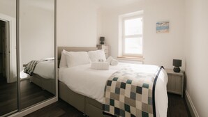 Un dormitorio moderno con un espejo grande, una cama con ropa de cama blanca, una colcha y una mesita de noche pequeña con una lámpara.