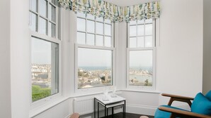 Habitación de esquina con tres ventanas amplias, una pequeña mesa con dos vasos y dos sillas azules.
