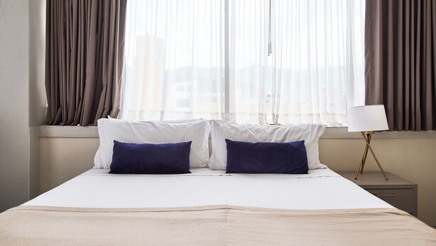 Een netjes opgemaakt bed met twee kussens, een nachtkastje met een lamp en een raam met gordijnen.