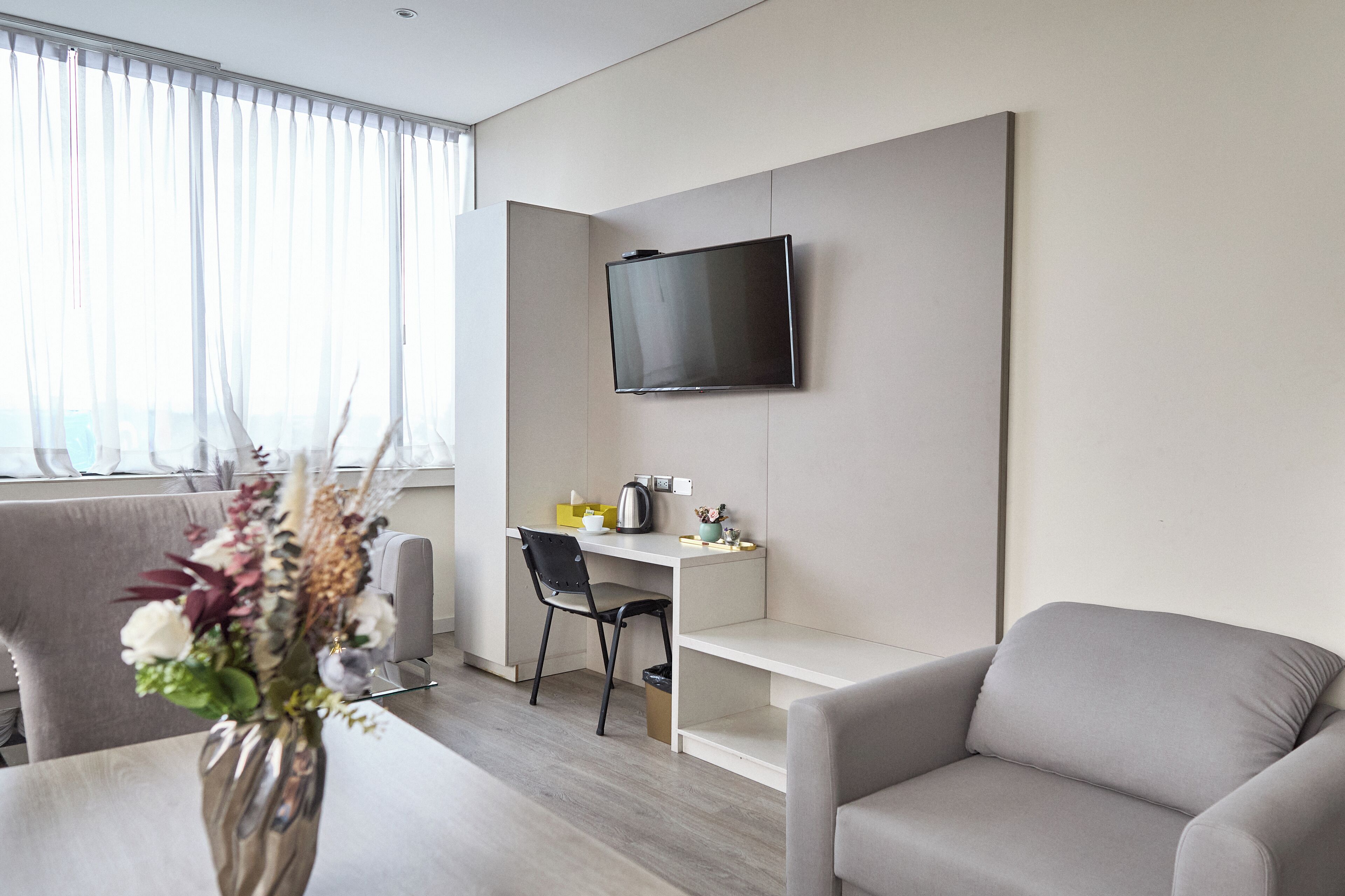 Una sala de estar moderna con un televisor de pantalla plana en la pared, un sofá gris, una mesa de comedor con un jarrón de flores y un sillón gris.