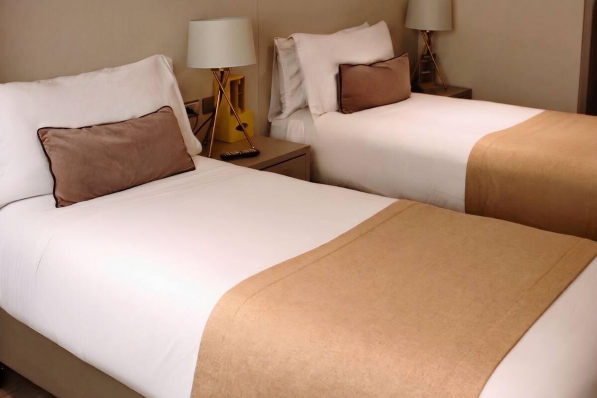 A hotel room with two beds, each with a brown throw blanket, white bedding, and bedside lamps.