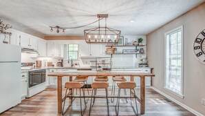 Una cocina moderna con armarios blancos, una isla de madera con taburetes de bar, una lámpara de araña y un reloj grande en la pared.