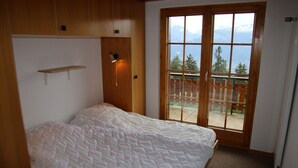 A bedroom with a bed, a wooden headboard, a small shelf, a window with a view of mountains and a balcony.