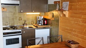 Een kleine keuken met een fornuis, spoelbak en koffiezetapparaat. Er staat een houten tafel met stoelen en een mand met fruit.
