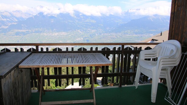 Een balkon met een houten tafel en een witte plastic stoel, met uitzicht op een bergachtig landschap, een vallei en een meer.