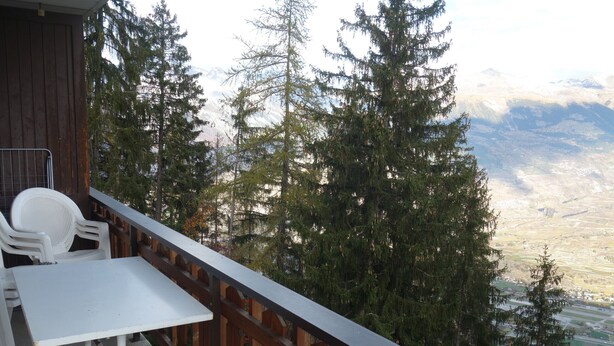 Un balcón con una mesa y sillas blancas, con vistas a un paisaje montañoso y árboles frondosos.