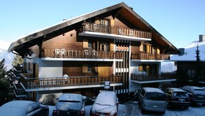 Una cabaña de madera de varios pisos con balcones, un telesilla y varios coches aparcados delante.