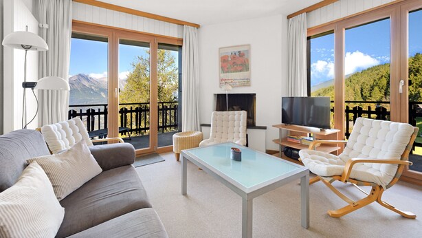 Ein geräumiges Wohnzimmer mit einer Couch, einem Glastisch, einem weißen Sessel und einem Fernseher. Große Fenster bieten Blick auf eine Berglandschaft.