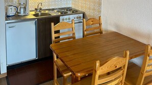 Cocina compacta con estufa, lavamanos y un refrigerador pequeño. Hay una mesa de comedor de madera con sillas y un cuadro en la pared.
