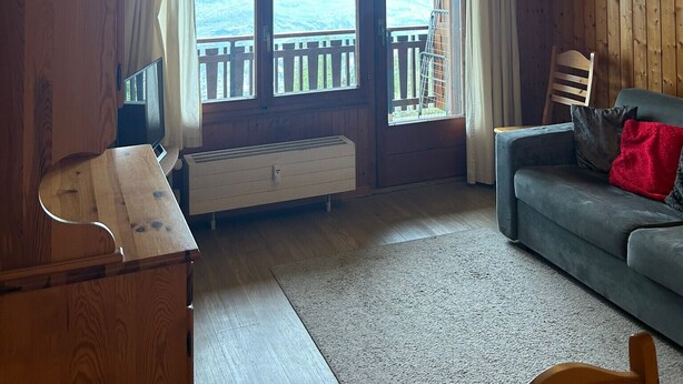 Una sala de estar acogedora con piso de madera, un sofá gris, un mueble de televisión de madera y un ventanal con vistas a un balcón.