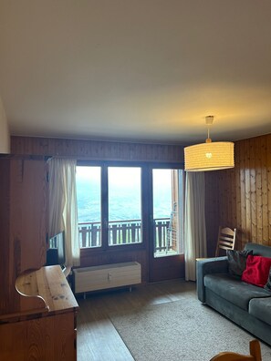 A cozy living room with a wooden floor, a grey sofa, a wooden TV stand, and a large window offering a view of a balcony. - Bellevue L 013 - PLEASANT studio 4 pers (Veysonnaz)