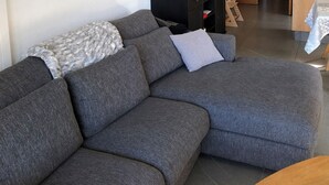 Een moderne woonkamer met een grijze modulaire bank, een witte pluizige kroonluchter, een klein houten bijzettafeltje en uitzicht op de aangrenzende eetruimte.