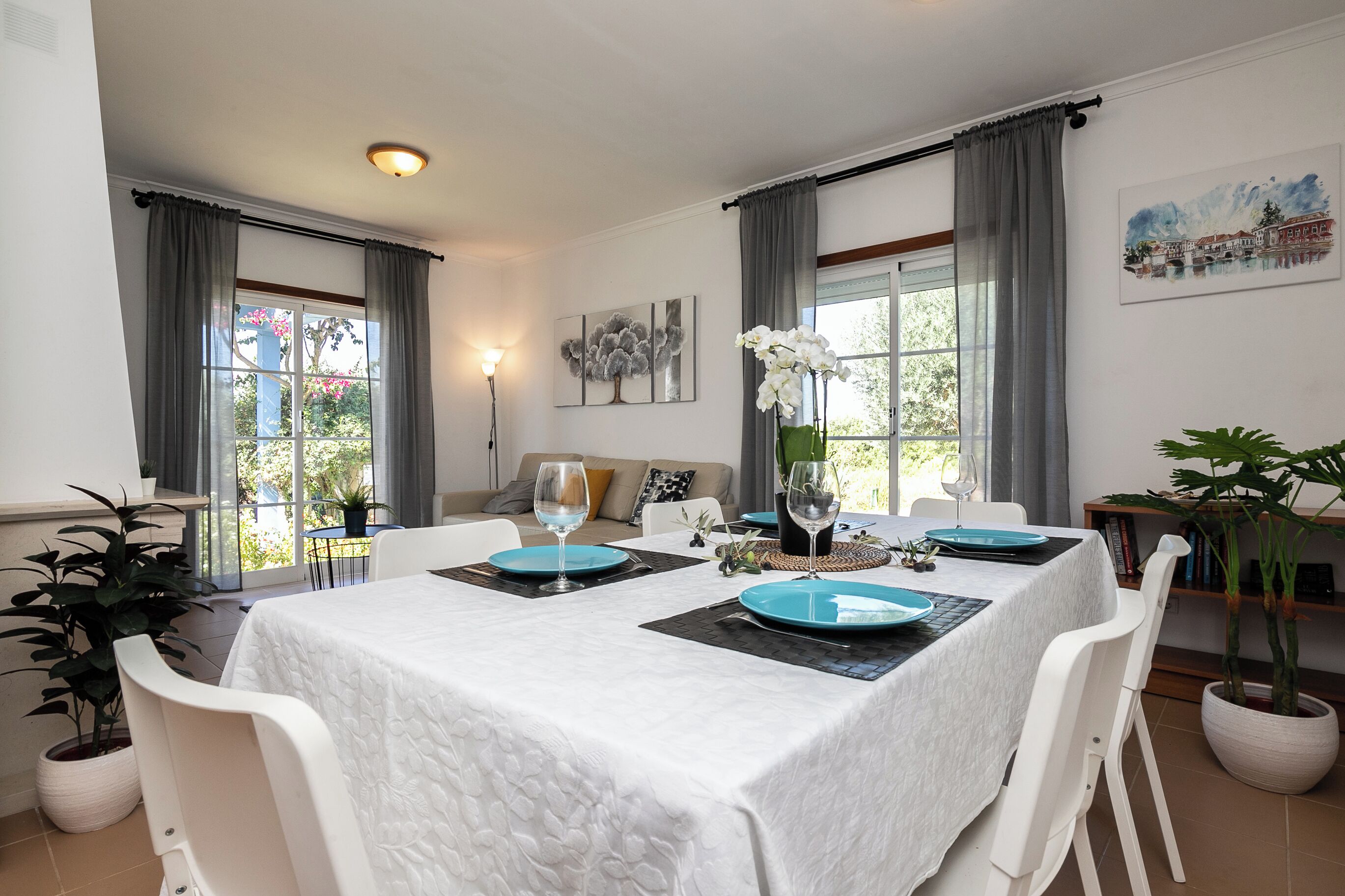 Un comedor con una mesa dispuesta para cuatro personas, sillas blancas, un mantel blanco y una planta en maceta.
