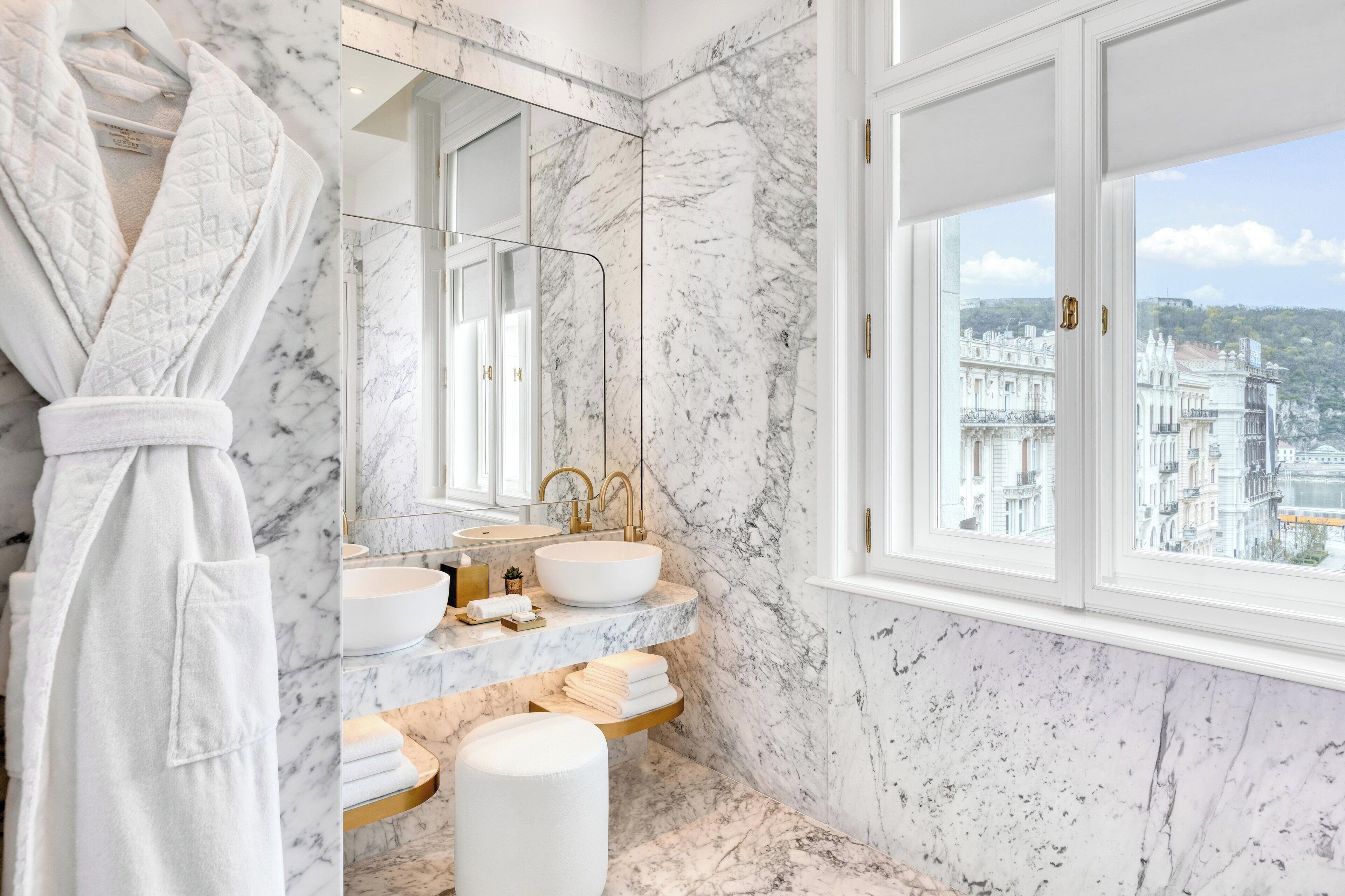 A bathroom with marble walls, a large window, a gold faucet, and a white bathrobe.