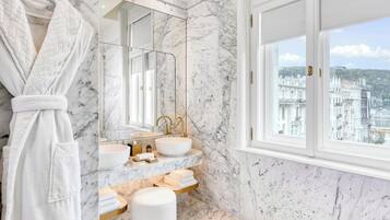 A bathroom with marble walls, a large window, a gold faucet, and a white bathrobe.
