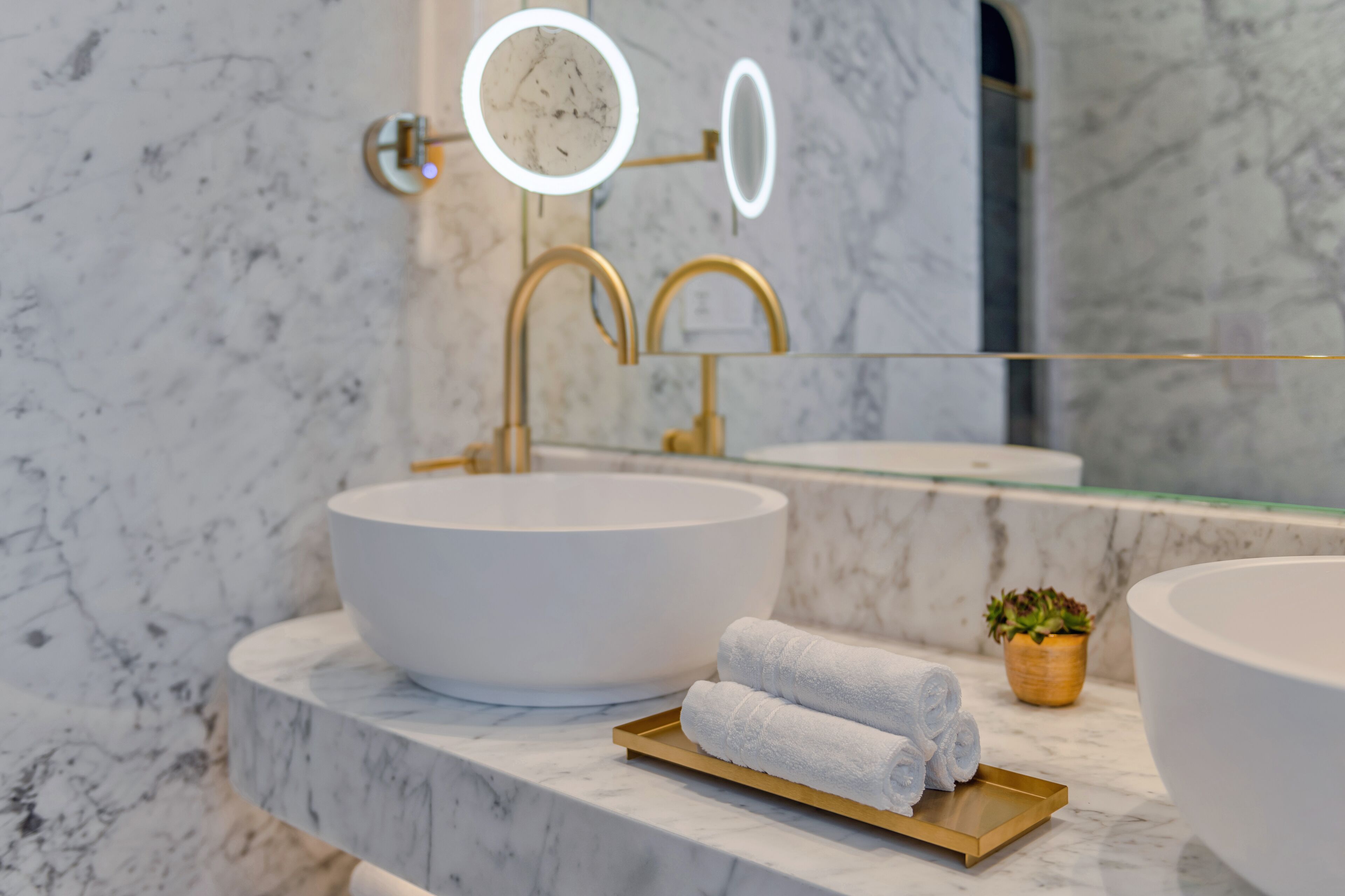 Une salle de bain avec un grand lavabo blanc, un robinet doré et un miroir doté de deux lampes. On y trouve deux serviettes enroulées sur un plateau et une petite plante en pot posée sur le comptoir.