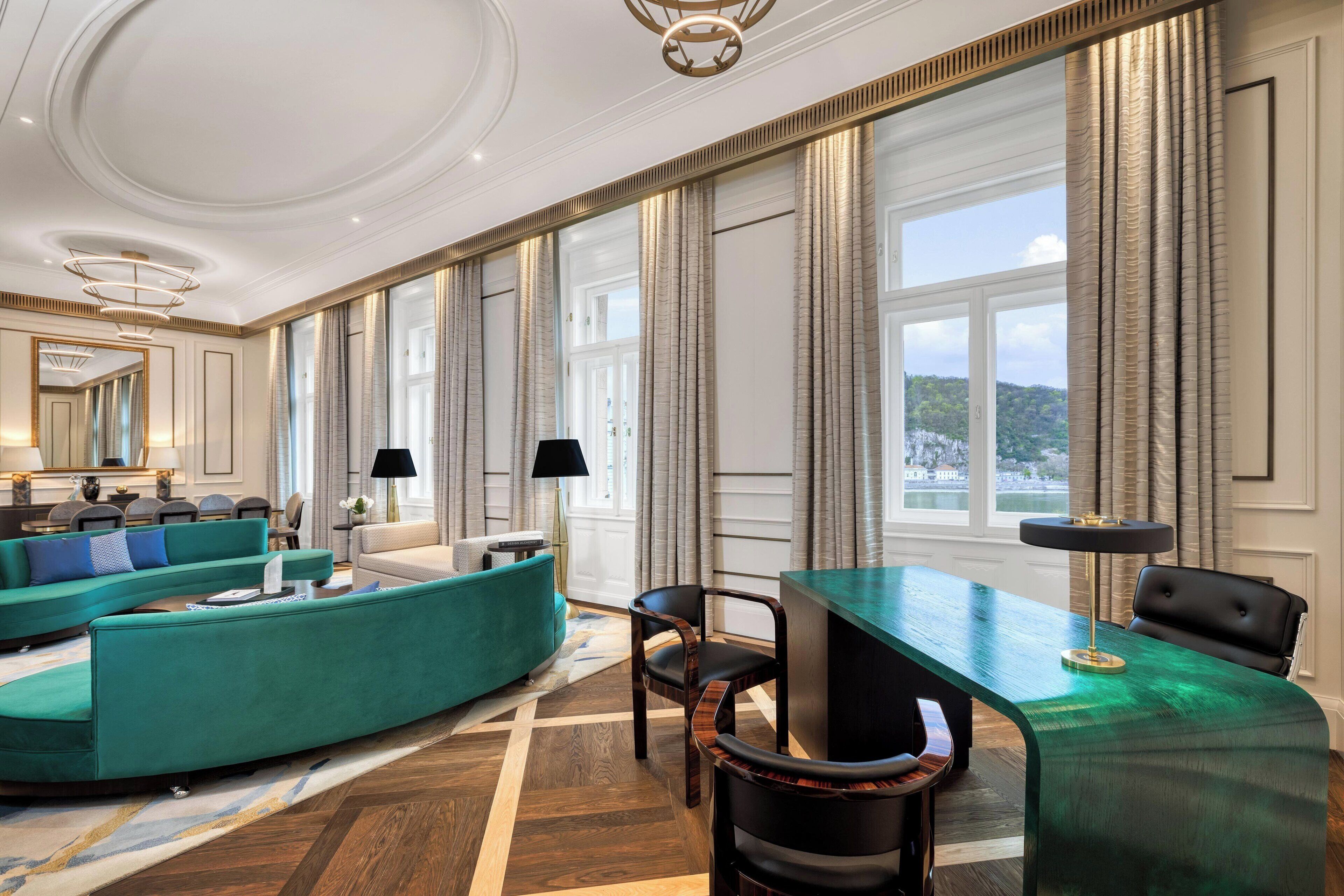 A spacious room with a curved teal sofa, a green desk, and a large window with curtains.