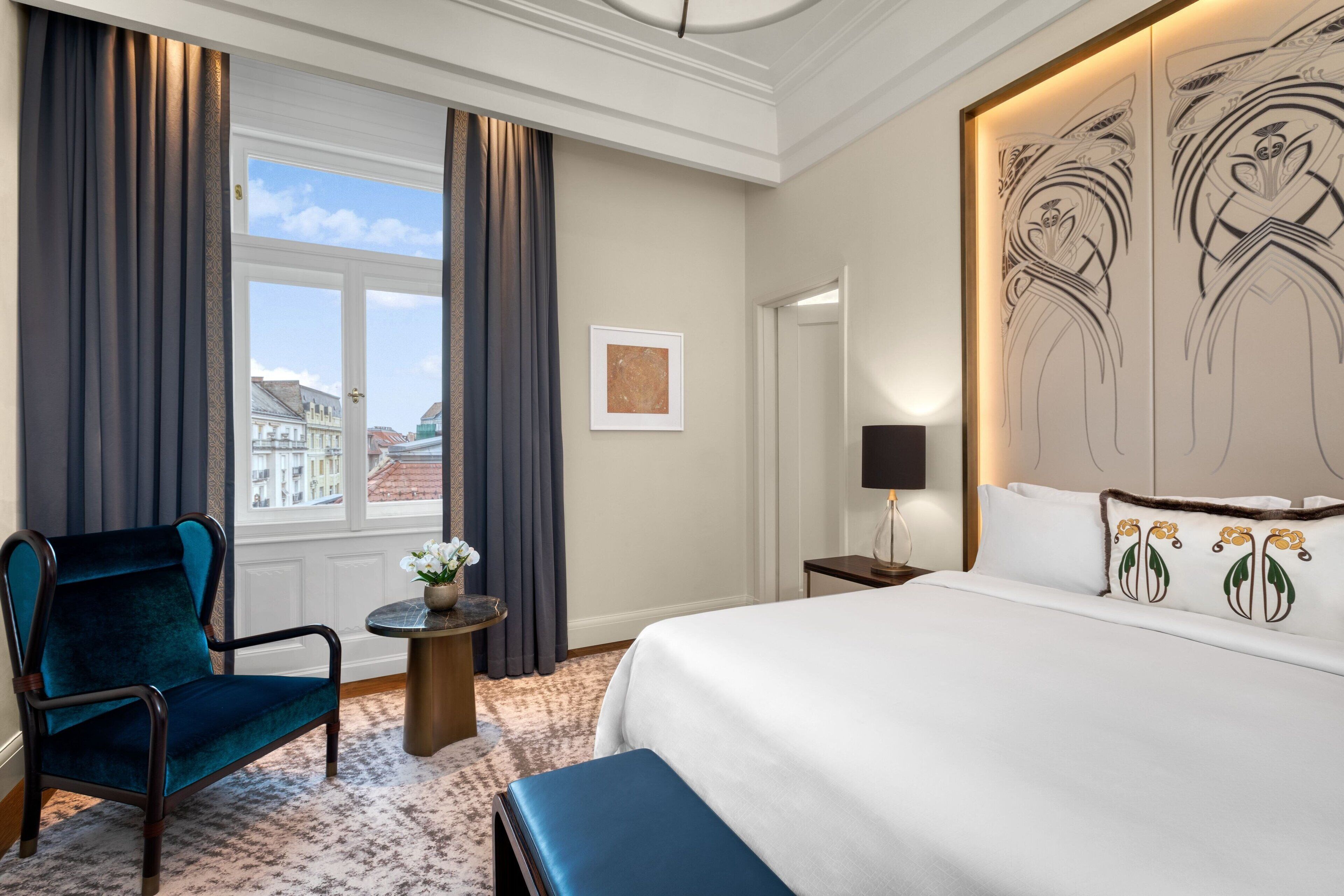Une chambre d’hôtel avec un grand lit, un fauteuil bleu foncé, une petite table avec un vase, et une vue sur les bâtiments par la fenêtre.