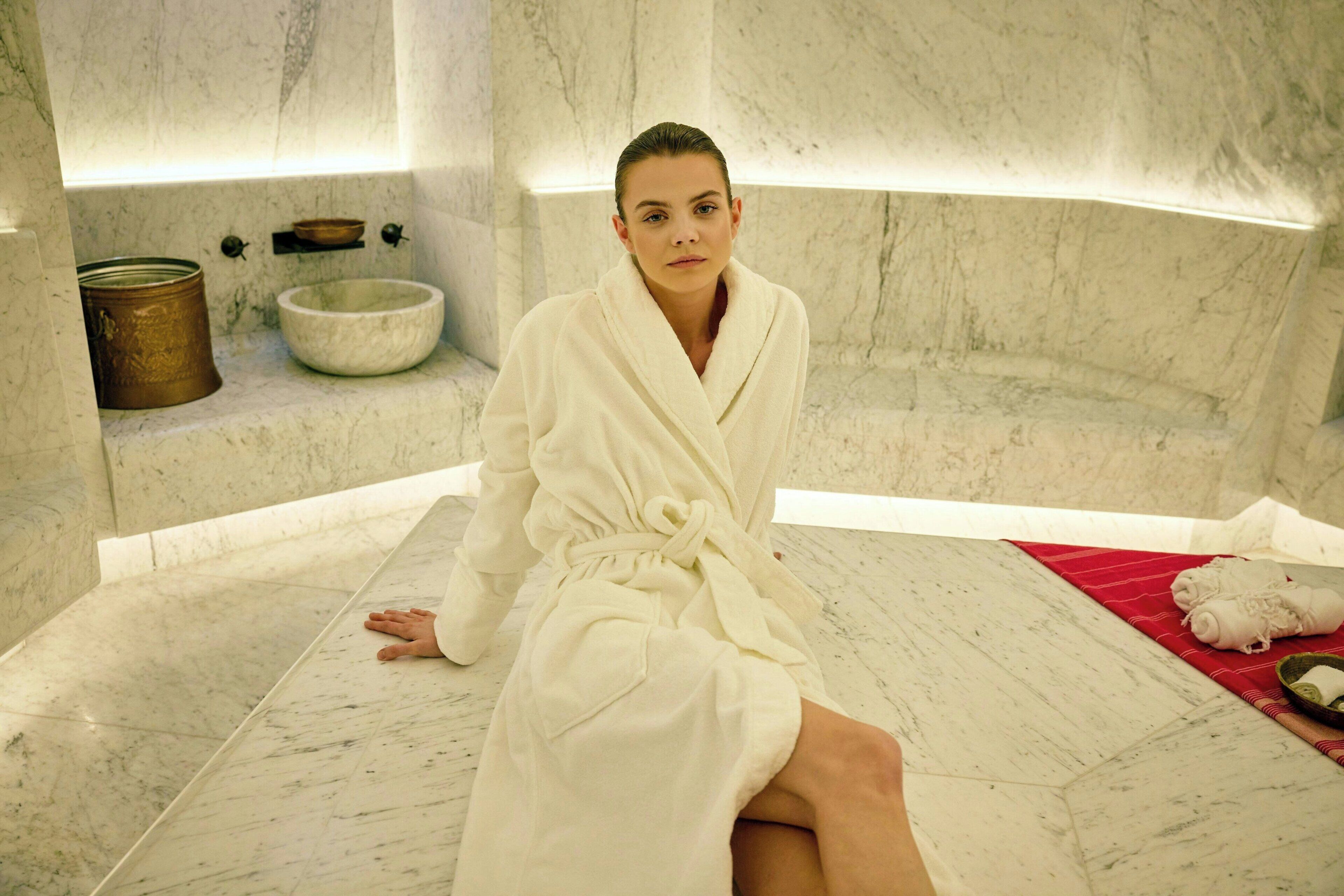 Henkilö istuu marmorilattialla spa-tyylisessä tilassa. Lähellä on suuri kulho ja kuparinen astia hyllyllä.