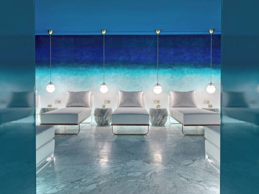 Un espacio de spa con cuatro zonas de estar acolchadas de color blanco, suelo de mármol e iluminación colgante.