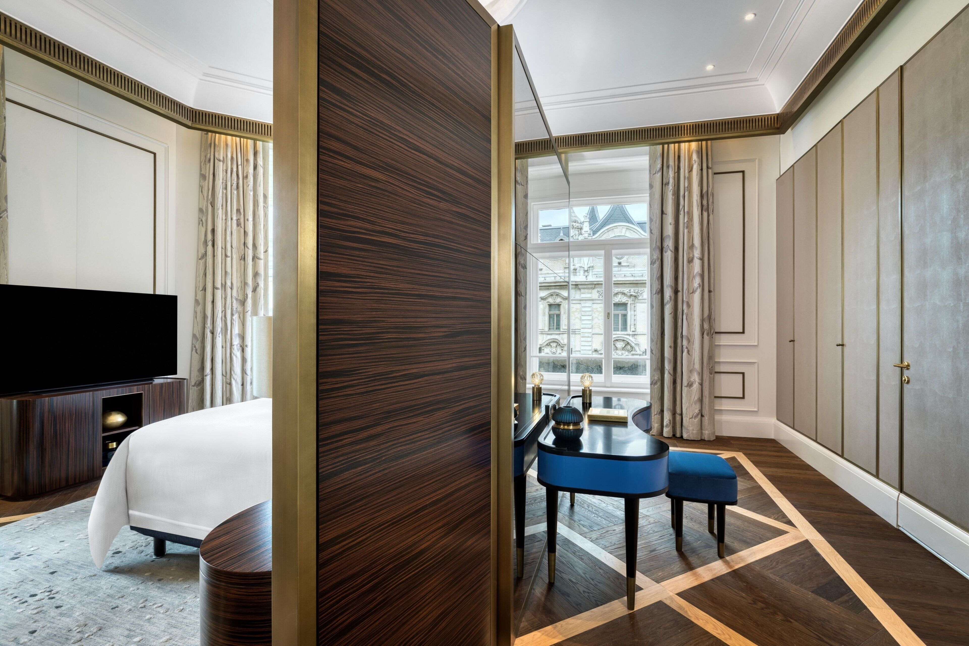 A modern hotel room with a large bed, a flat-screen TV, a dining area with a blue table and chairs, and a view of a historic building through large windows.