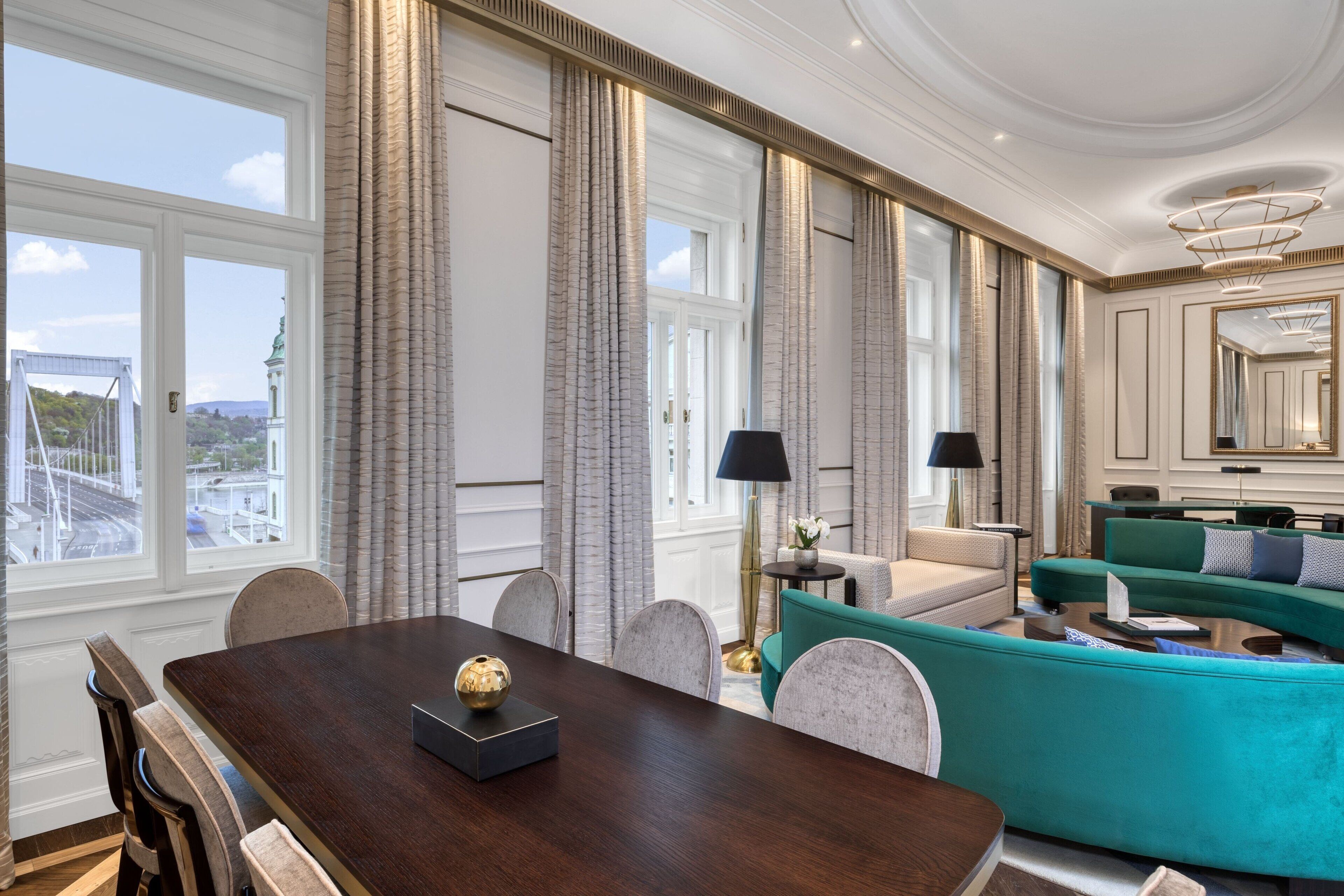 A luxurious room with a dining table, teal sofa, and large windows offering a city view.
