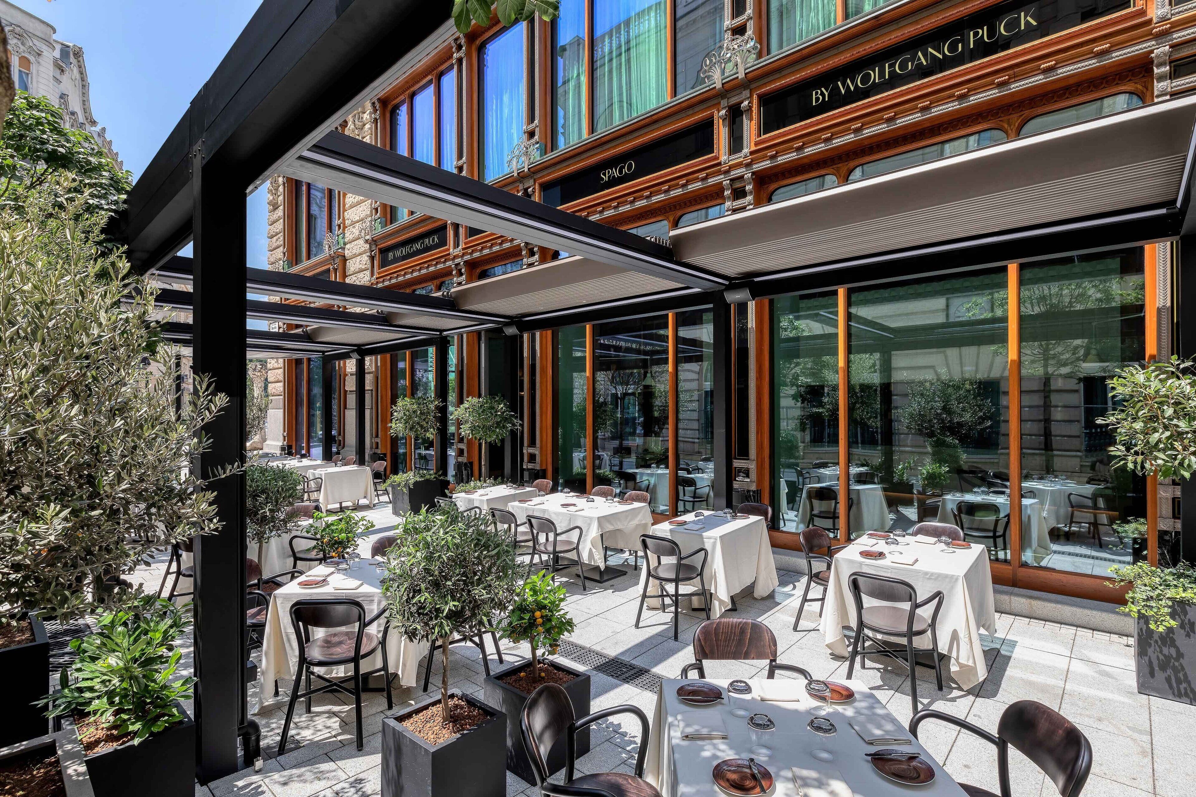 An outdoor dining area with tables and chairs, surrounded by potted plants and large windows.