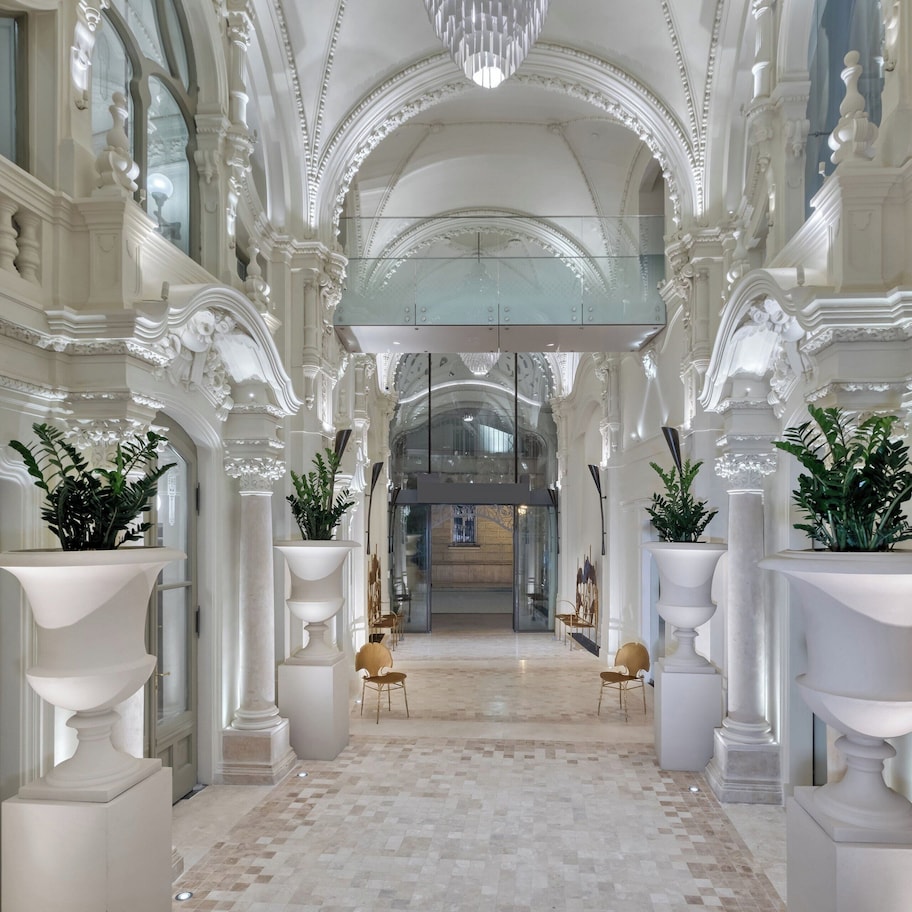 An ornate hallway with white columns, large planters, and a grand staircase.
