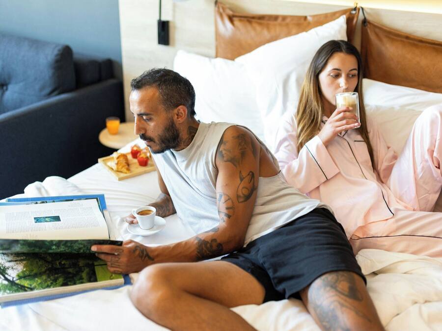 Un dormitorio con una cama, almohadas, una mesita de noche con un libro y una taza, y un aparador con un frutero.